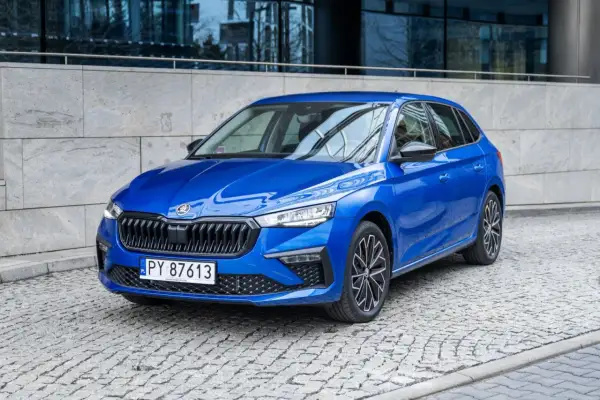 Błękitna Skoda Scala 2024 z czarnymi felgami stoi na brukowanej ulicy przed nowoczesnym budynkiem.