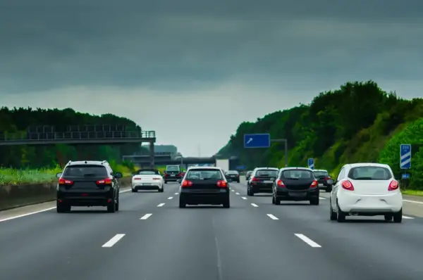 Samochody jadące drogą szybkiego ruchu pod pochmurnym niebem. W oddali widać zielone drzewa i wiadukt.