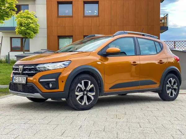 Pomarańczowy Dacia Sandero Stepway na tle nowoczesnego budynku. Sprawdź atrakcyjne dacia sandero - ceny.
