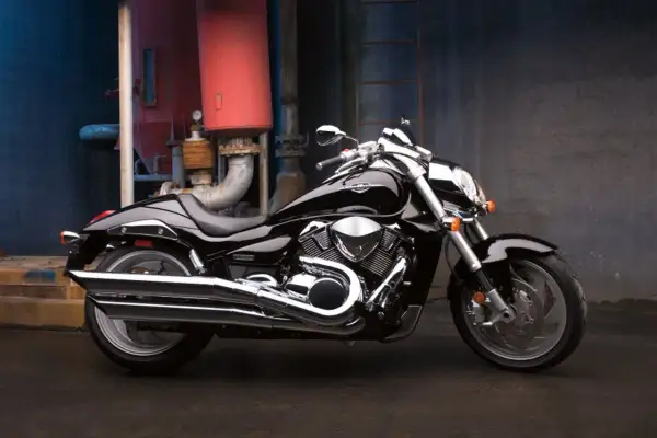 Błyszczący, czarny Suzuki Intruder 1800 z chromowanymi elementami, zaparkowany na tle industrialnej ściany z rurami.
